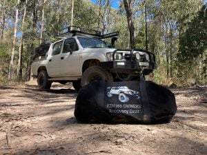 White 4WD vehicle equipped with off-road modifications such as a roof rack, bull bar, and spotlights, parked in a forest setting. In the foreground, a large black bag labeled 'SZN 185 OWNERS RECOVERY GEAR' is placed on the ground. This Explore Off Road scene highlights essential recovery gear and accessories for 4WD enthusiasts, emphasizing the importance of being prepared for off-road adventures in Australia.
