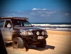 4WD vehicle driving on a sandy beach with the ocean in the background, equipped with off-road modifications including a roof rack, bull bar, and spotlights. This Explore Off Road vehicle demonstrates the capabilities of off-road accessories in a beach environment, ideal for adventure seekers and 4WD enthusiasts in Australia.